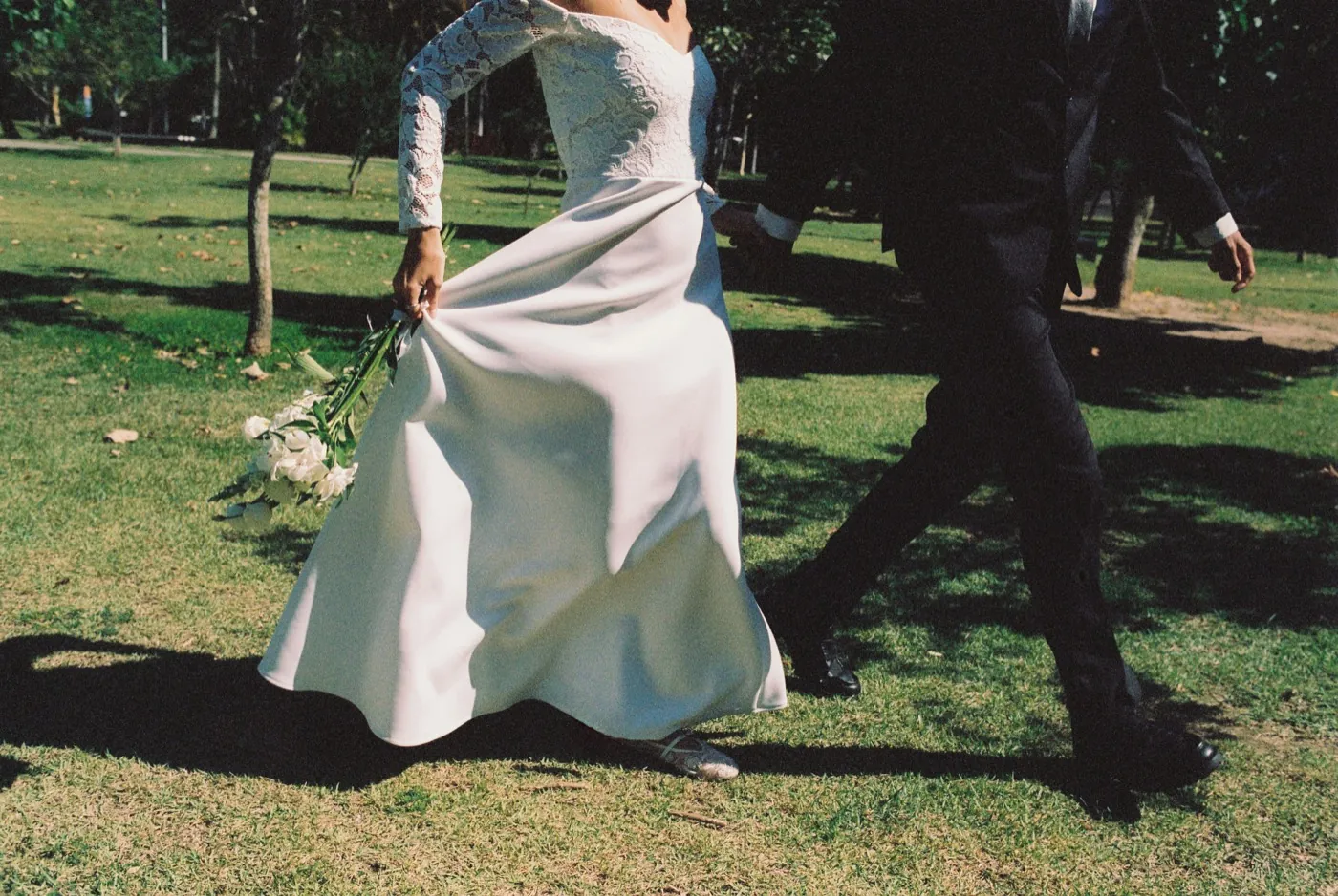 A couple walking across sunlit grass, bride lifting her dress