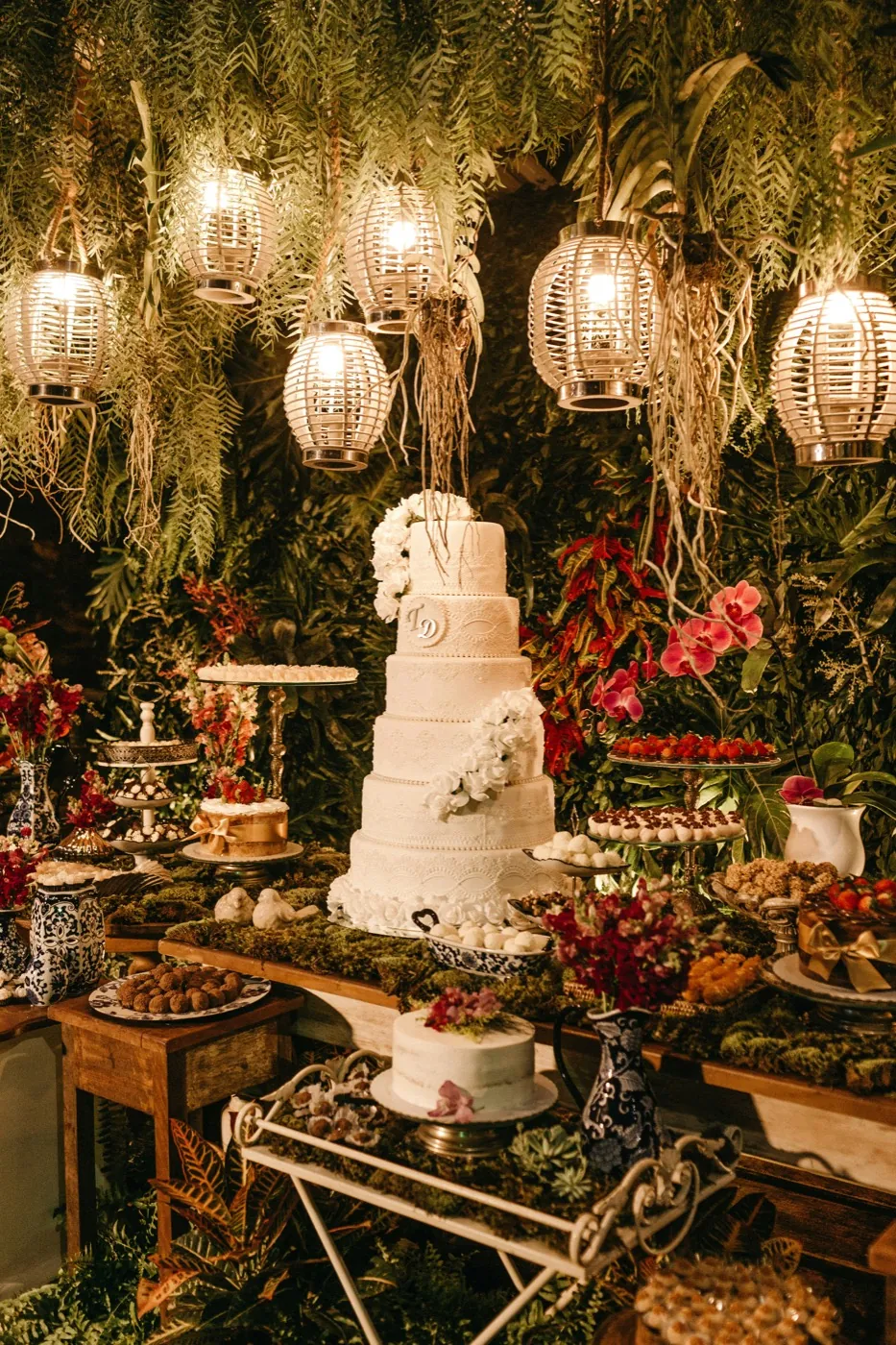 A dessert table under hanging lanterns and ferns, tiered lace cake