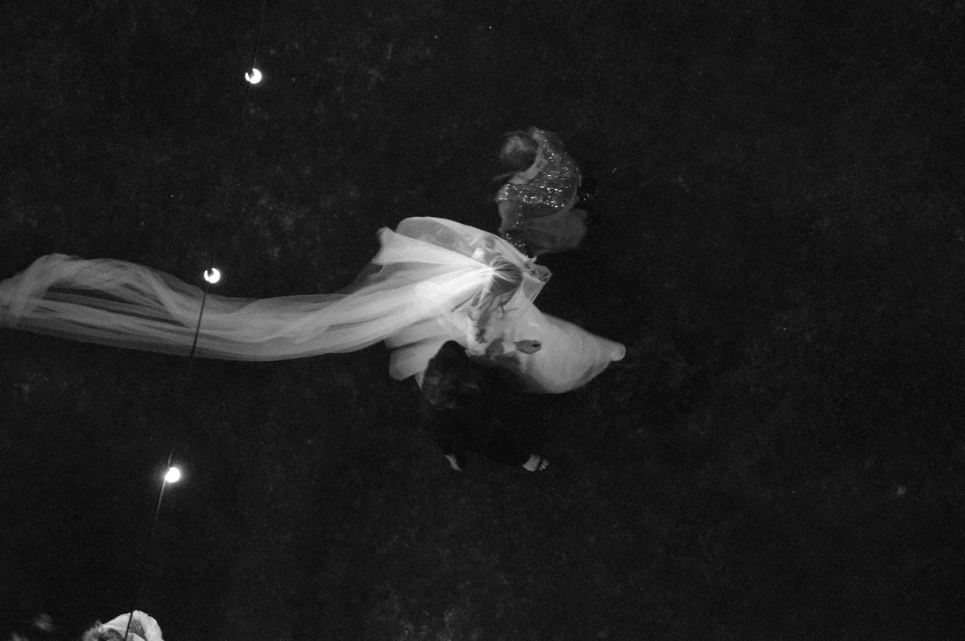 Black and white, seen from above: a couple dancing at night, her long veil flying past a string of lights