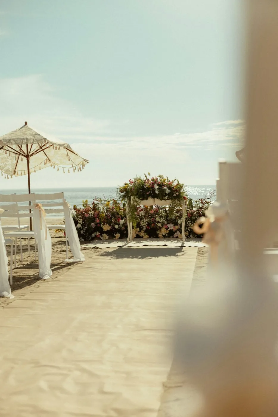 A beach ceremony aisle with flowers and a fringed parasol by the sea