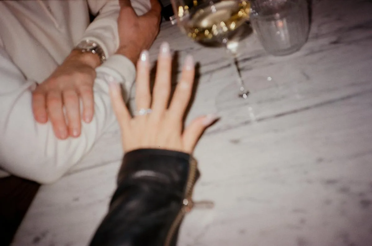 A hand with a new engagement ring resting on a marble table, a partner's hand and a glass of wine beside it